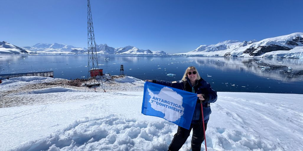 Tracy holding an "Antarctica 7th Continent" flag on the snow, with a red base, antenna tower and a wide ice-filled bay behind