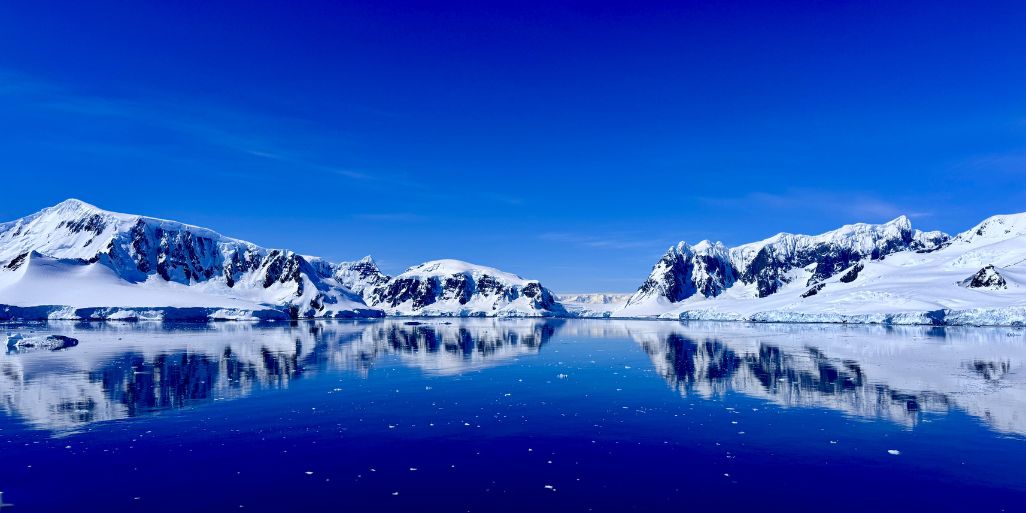 A panorama of snow-covered mountains reflected in a deep blue, mirror-calm Antarctic bay under a clear sky