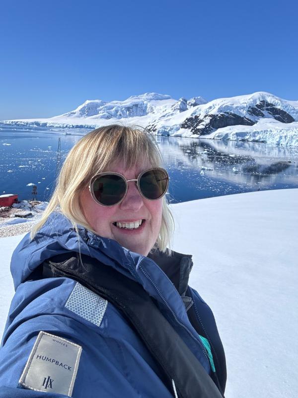 Tracy Collins in an HX expedition parka smiling in a selfie, with snow-covered mountains and calm water behind in Antarctica