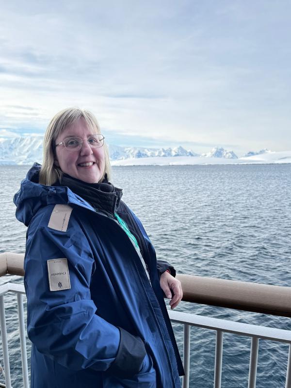 Tracy Collins in an HX expedition parka smiling in a selfie, with snow-covered mountains and calm water behind in Antarctica