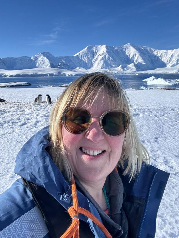 Tracy Collins in sunglasses taking a selfie, with Gentoo penguins, snowy ground and mountains behind in Antarctica