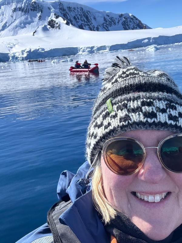 Tracy Collins in a bobble hat taking a selfie, with a zodiac boat and kayakers on calm water and snowy mountains behind