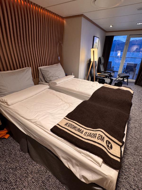 Twin beds in a balcony cabin on MS Roald Amundsen, with a branded wool blanket folded across the foot of the bed and a sea view through the window